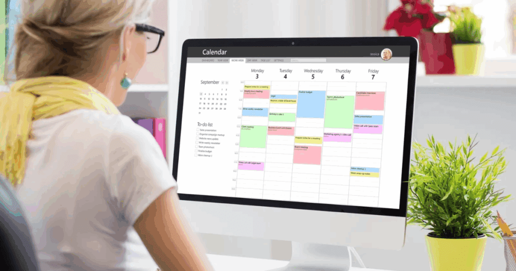 Woman looking at a desktop computer screen displaying a colorful weekly calendar and to-do list, with a potted plant on the desk beside her.