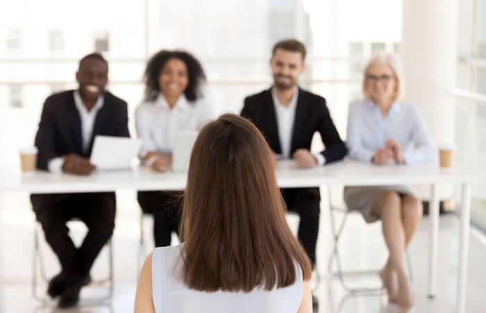 A woman sits facing a panel of four smiling interviewers across a table in a bright, modern office setting.